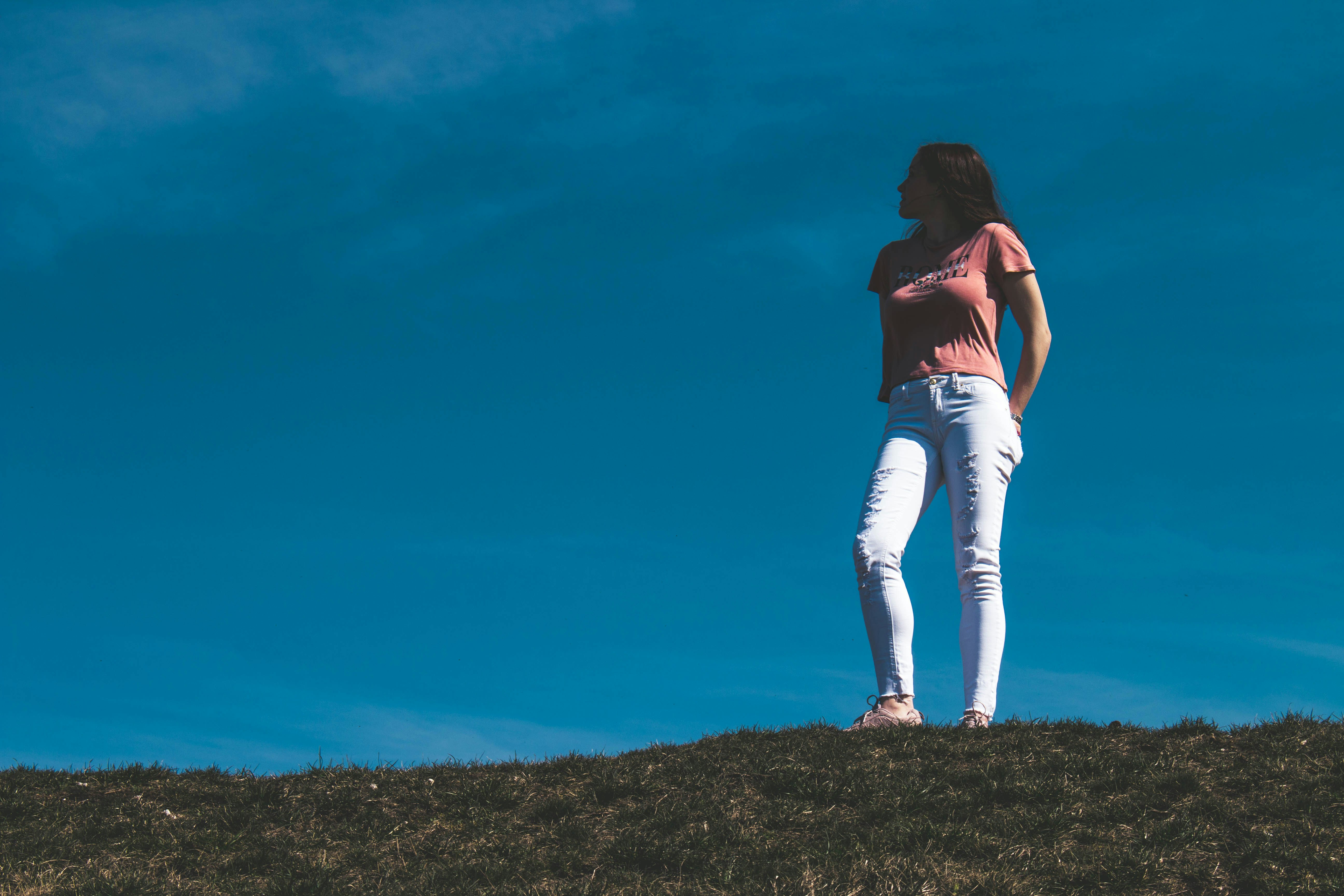 woman in pink t-shirt and white jeans standing on hill under blue sky